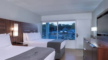 A hotel room with two beds, a TV, a desk, and a large window with a view of a parking lot and buildings.