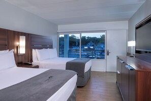 A hotel room with two beds, a TV, a desk, and a large window with a view of a parking lot and buildings. - Crosswinds Rehoboth  (Rehoboth Beach)