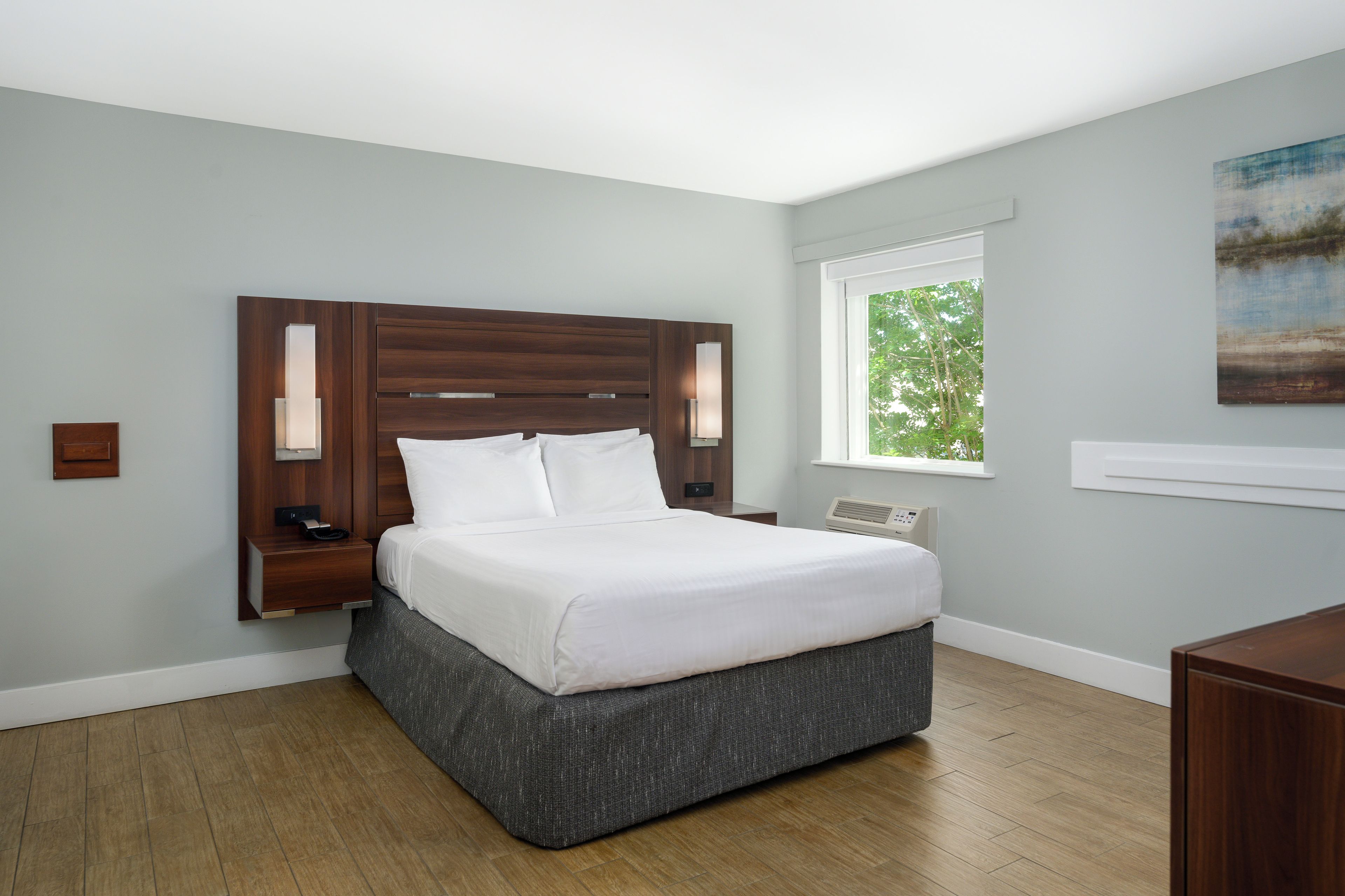 Une chambre moderne avec un grand lit, des tables de chevet, une commode, une fenêtre donnant sur des arbres et un tableau au mur.