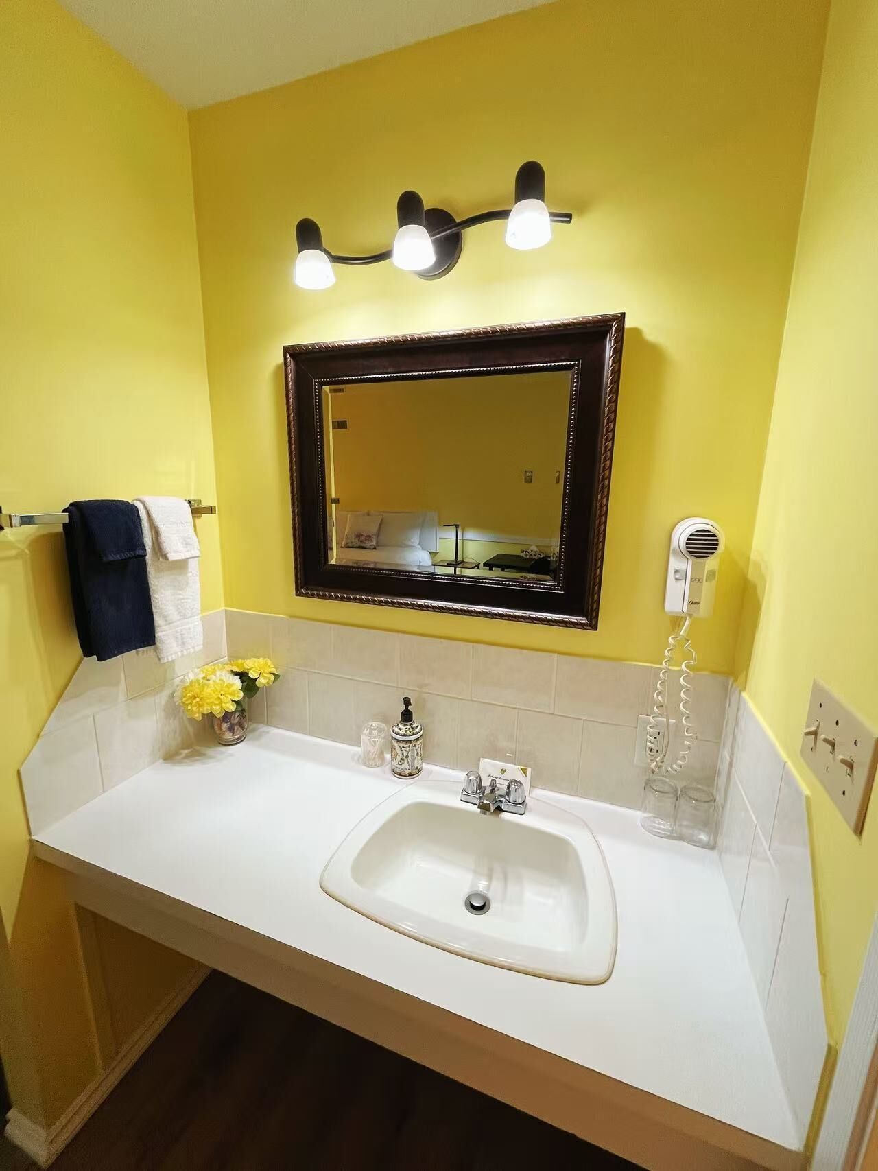 Een badkamer met een wit wastafelmeubel, gele wanden, een spiegel met verlichting en een handdoekhouder.