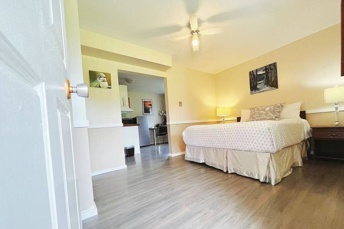 A bedroom with a bed, bedside tables, a desk, and a chair. There is a door leading to a kitchen area.
