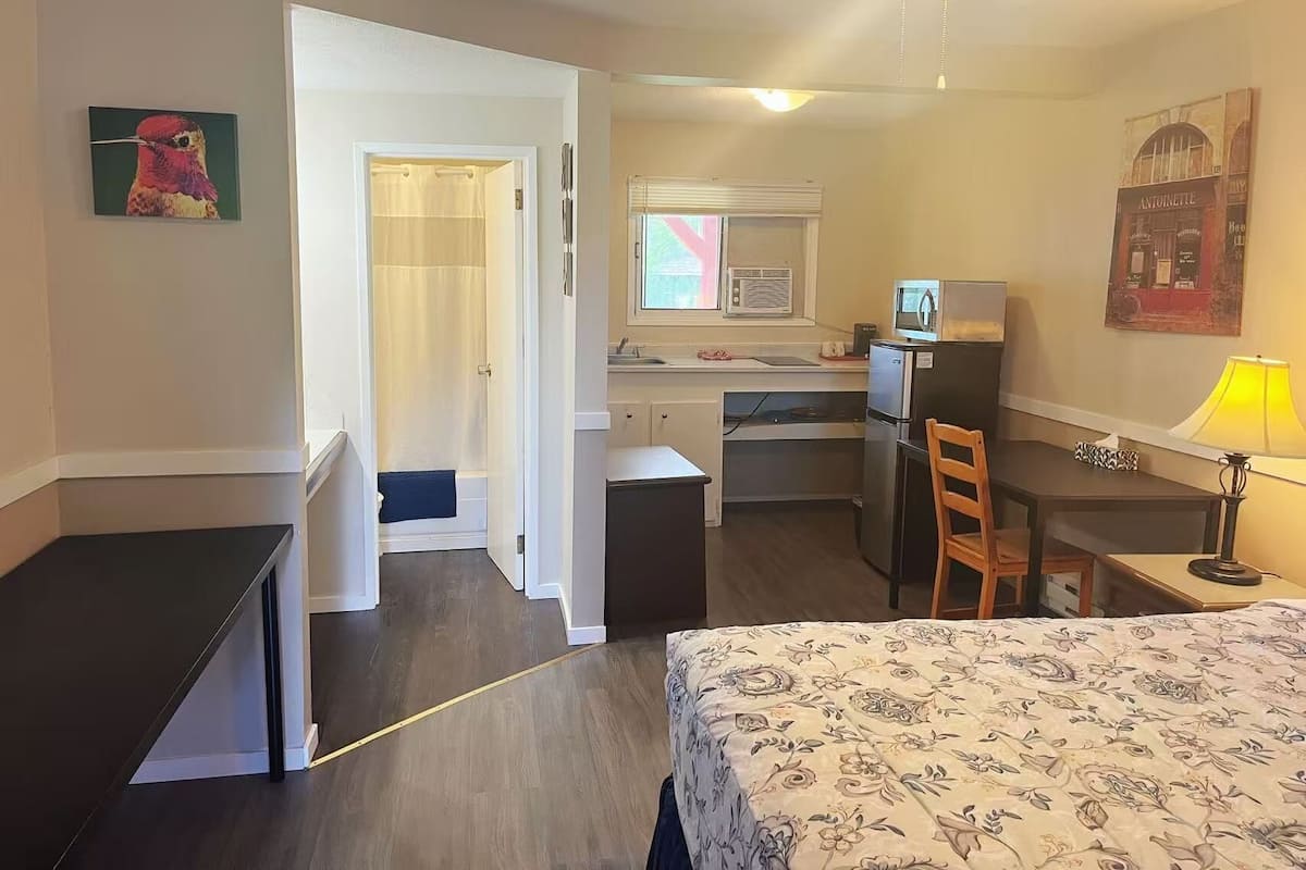 A compact hotel room with a bed, a desk, a chair, a small kitchenette, and a bathroom.