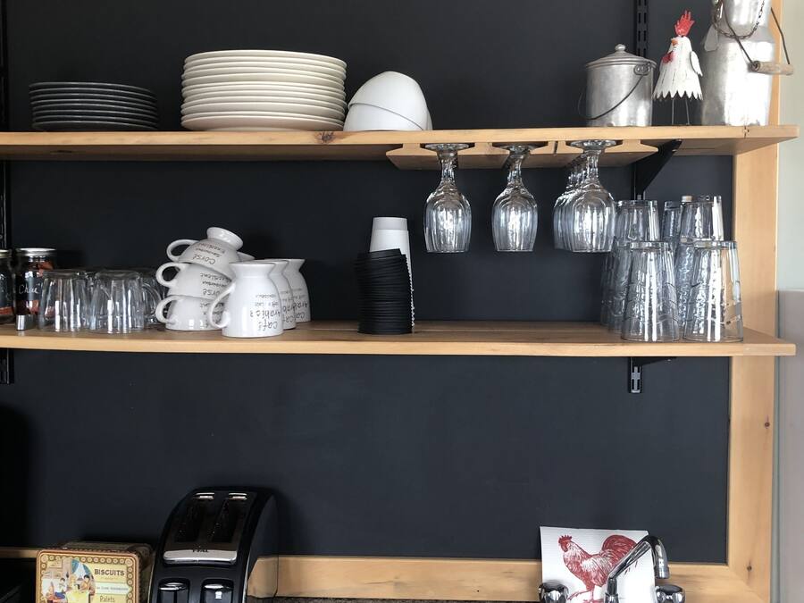 A kitchen with wooden shelves holding plates, bowls, and glasses. There is a toaster, a sink, and a cloth on the counter.