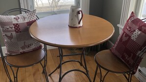 A dining area with a round wooden table, two metal chairs with patterned cushions, and a white pitcher on the table. - Auberge d'Anjou-Sūrya Café (Petit-Rocher)