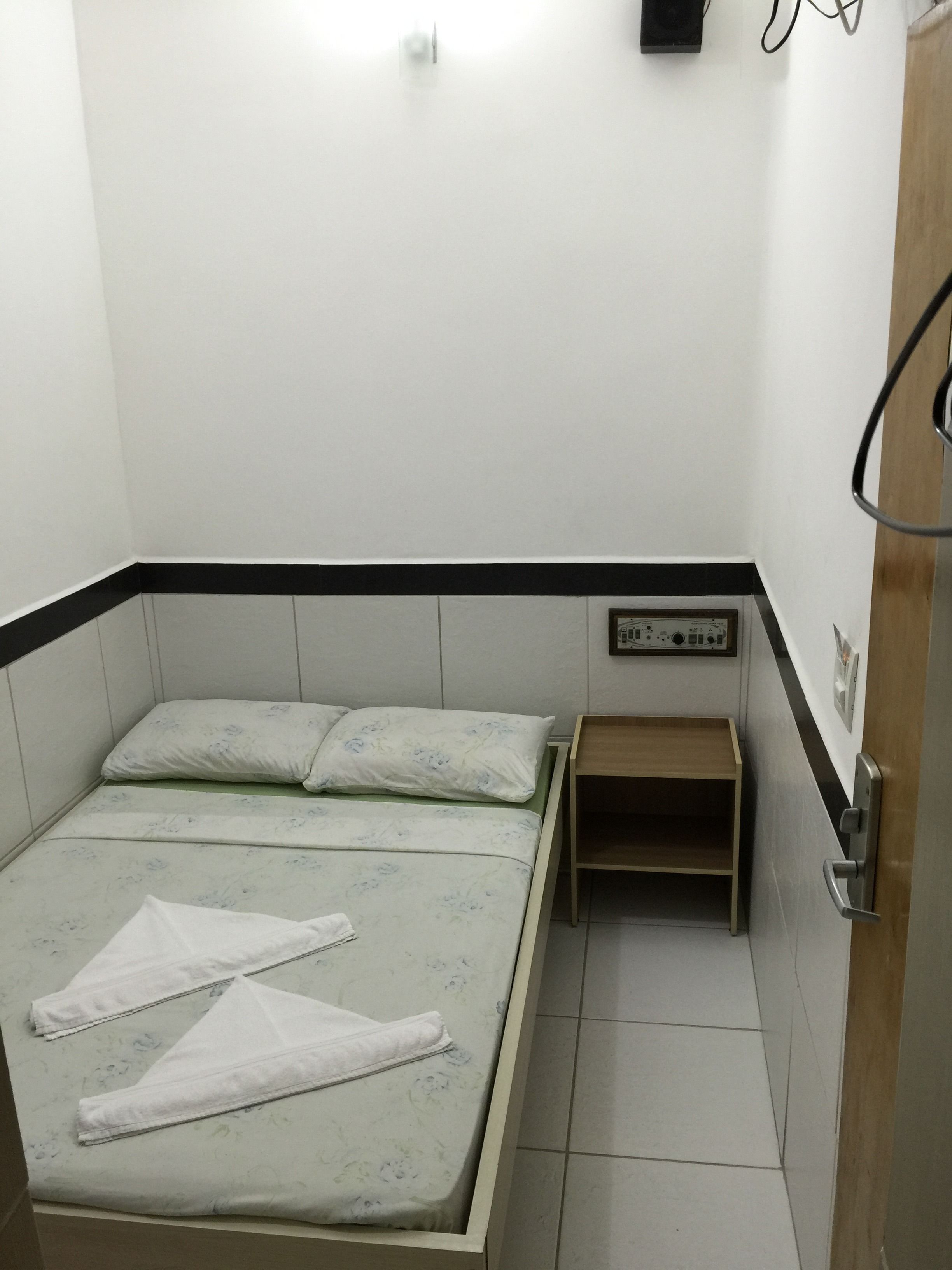 A small, clean room with a bed, a nightstand, and a wall-mounted control panel.