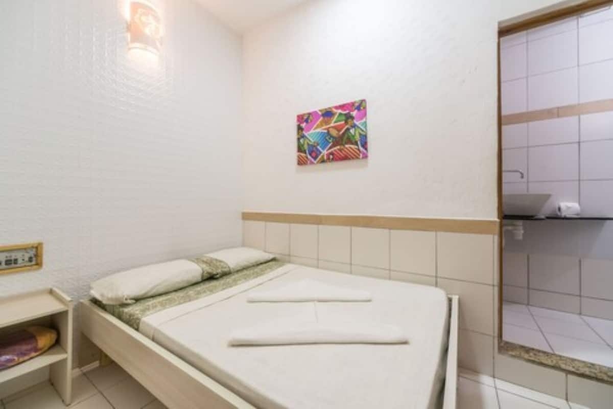 A hotel room with a bed, a small bedside table, a wall-mounted light, and a colorful artwork above the bed.
