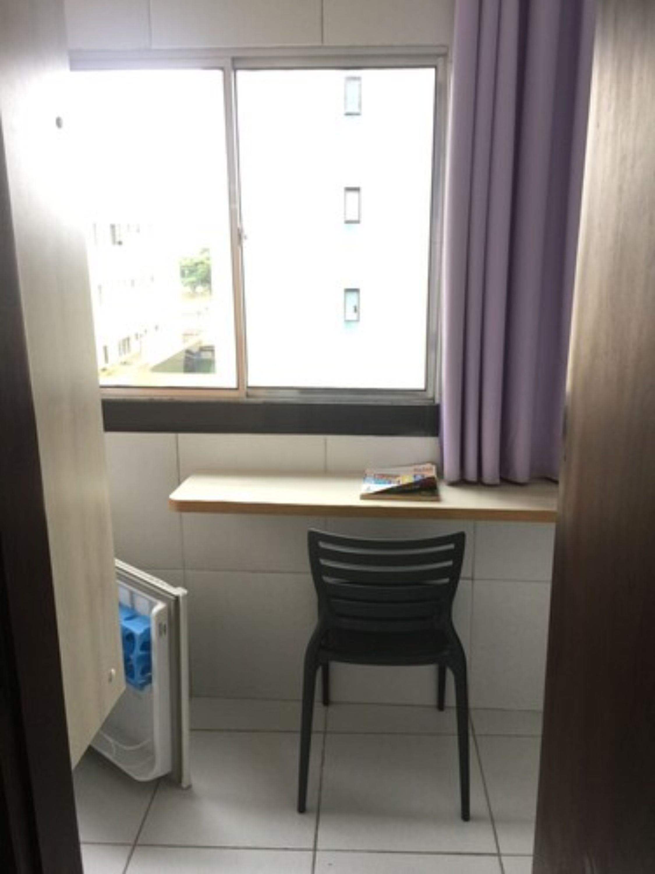 Une petite pièce avec une fenêtre, une chaise et une étagère sur laquelle se trouve un livre.