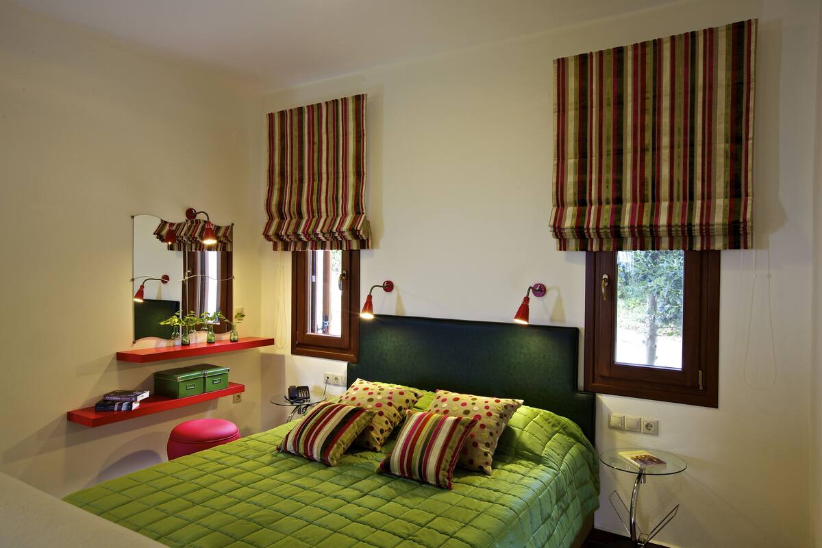 A bedroom with a green quilted bed, striped curtains, a mirror, and a small table with a book.