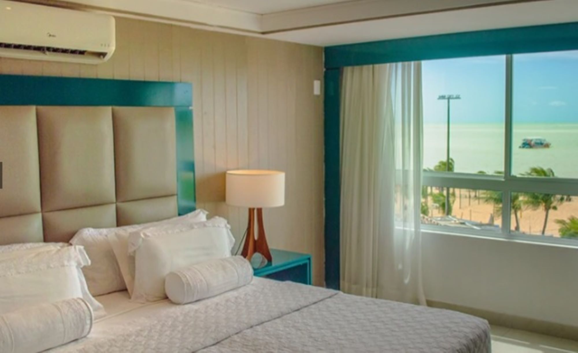 A hotel room with a bed, a nightstand, a lamp, and a view of the beach.