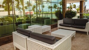 Un área de descanso moderna con mobiliario de mimbre, amplias ventanas que ofrecen vistas a palmeras y a una playa.