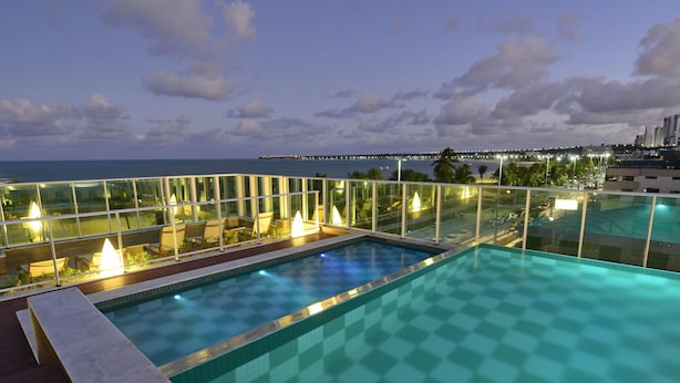 Una piscina infinita en la azotea con barandilla de cristal, vista al océano y luces de la ciudad a lo lejos.