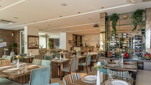Amplia sala de comedor de hotel con varias mesas dispuestas para comer, un área de bar con una bodega de vinos y un mostrador de recepción al fondo.