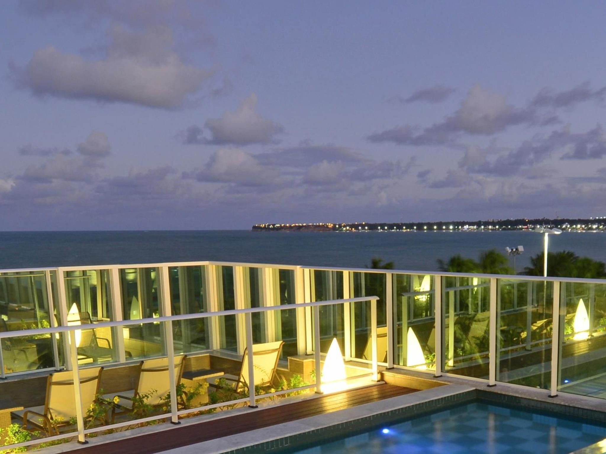 Un área de piscina en la azotea con sillones de descanso y vistas al océano y un muelle distante.