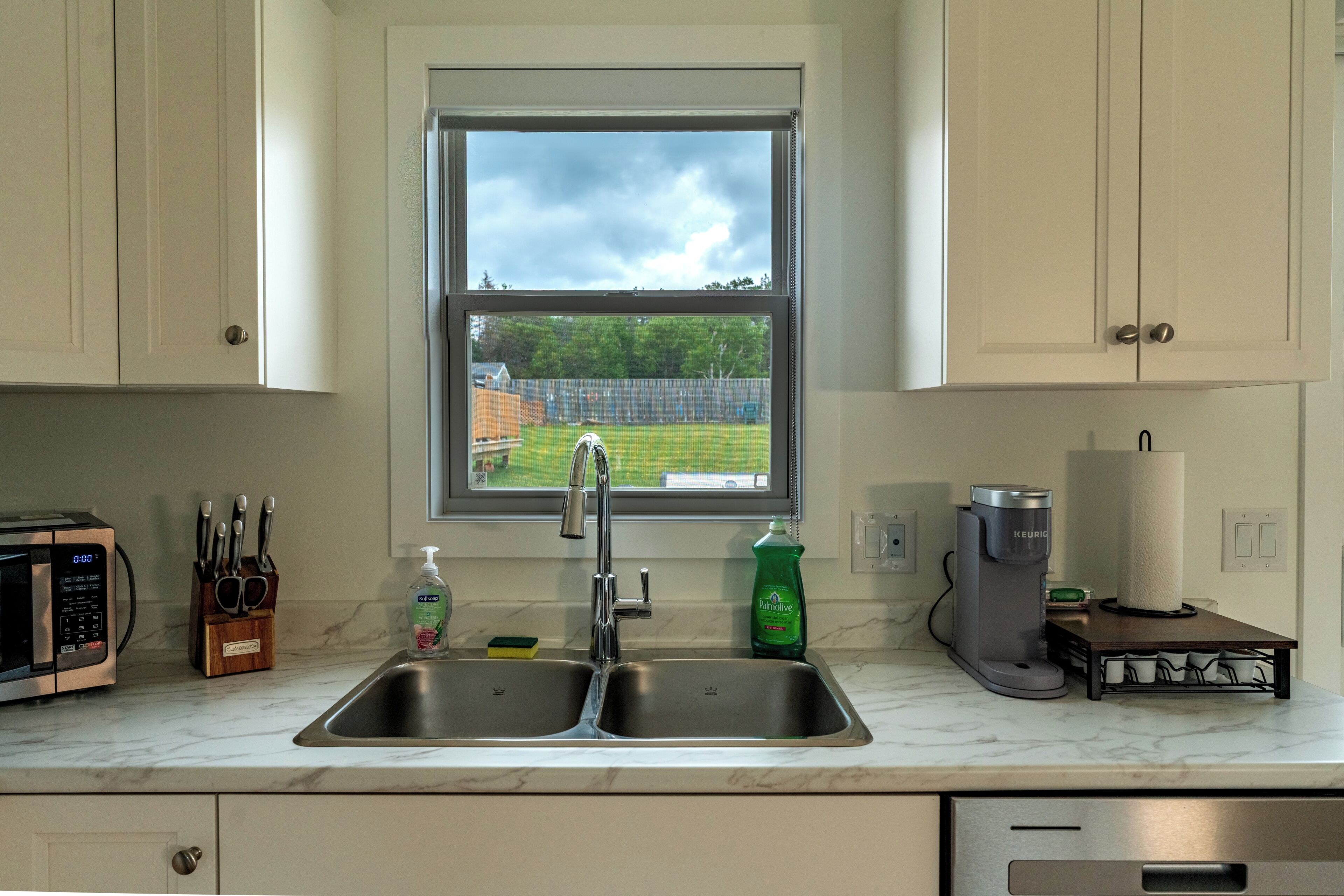 Una cocina con un lavamanos doble, una ventana, un microondas, una cafetera y un bloque de cuchillos.