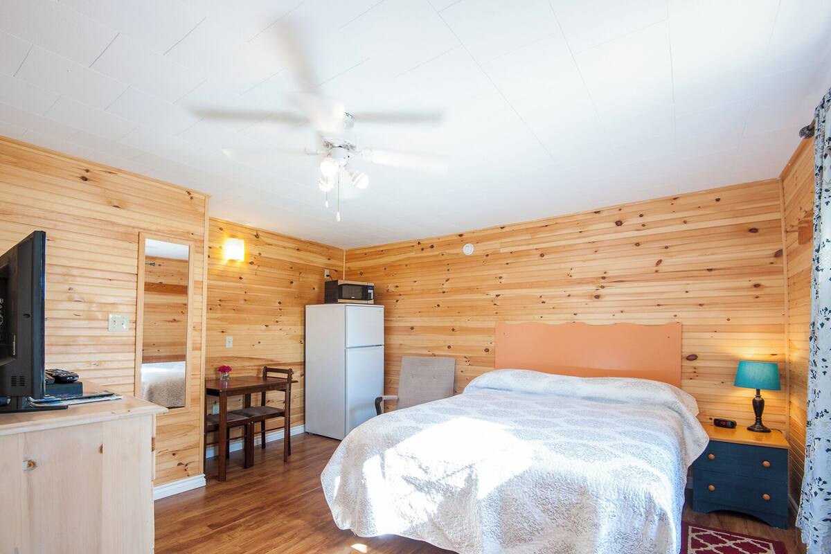 A wooden-paneled room with a bed, a desk, a chair, a television, a refrigerator, and a ceiling fan.