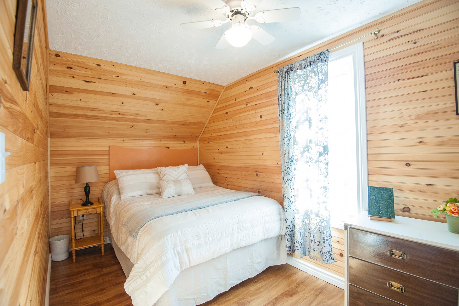 A bedroom with a wooden ceiling, a bed with white bedding, a nightstand, a lamp, a dresser, and a window with patterned curtains.