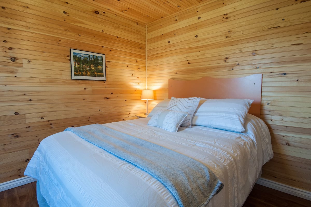 A wooden-paneled room with a bed, a nightstand, and a wall-mounted lamp.