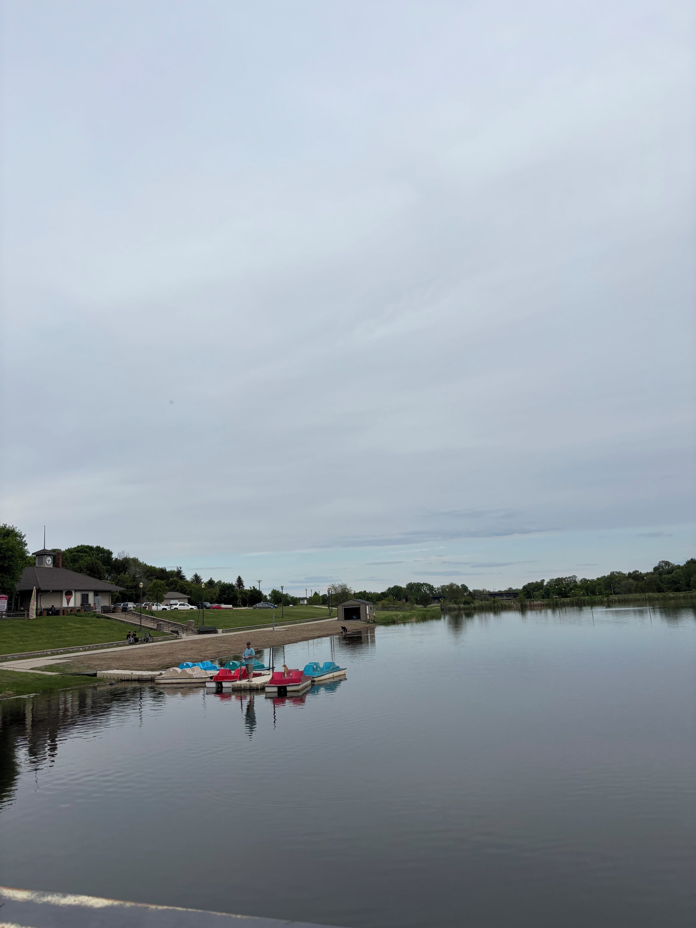 Un lac paisible, avec des bateaux amarrés sur la rive, un petit bâtiment et une zone herbeuse avec un sentier menant à l’eau.