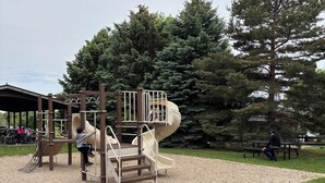 Un parque infantil con tobogán y estructuras para escalar, rodeado de árboles y una zona de picnic.