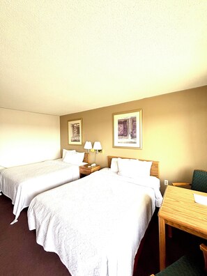 A hotel room with two single beds, a wooden nightstand with lamps, a wooden desk, and two framed pictures on the wall. - Dakota Inn (Huron)