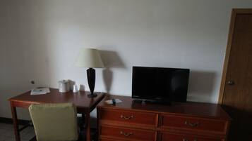 A hotel room with a wooden desk, a television, a lamp, a chair, and a wooden cabinet with drawers.