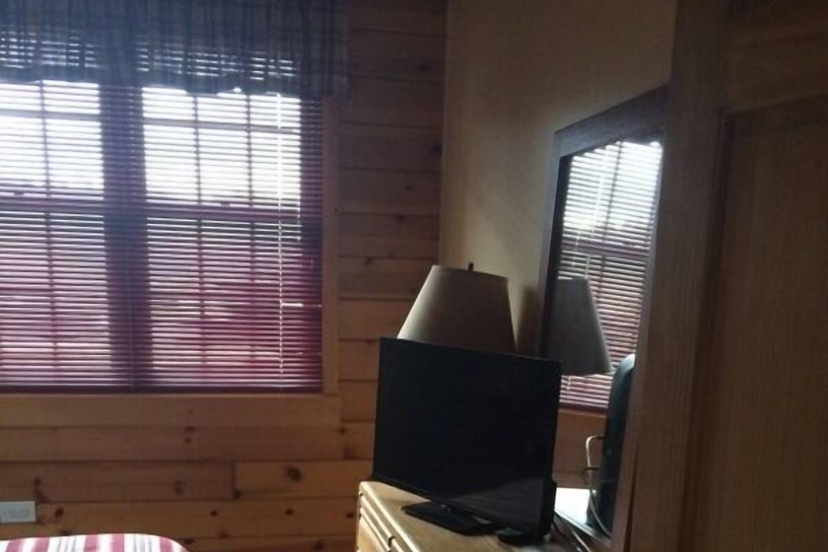 Une chambre à coucher avec un lit, une commode, un téléviseur et une fenêtre avec des stores.