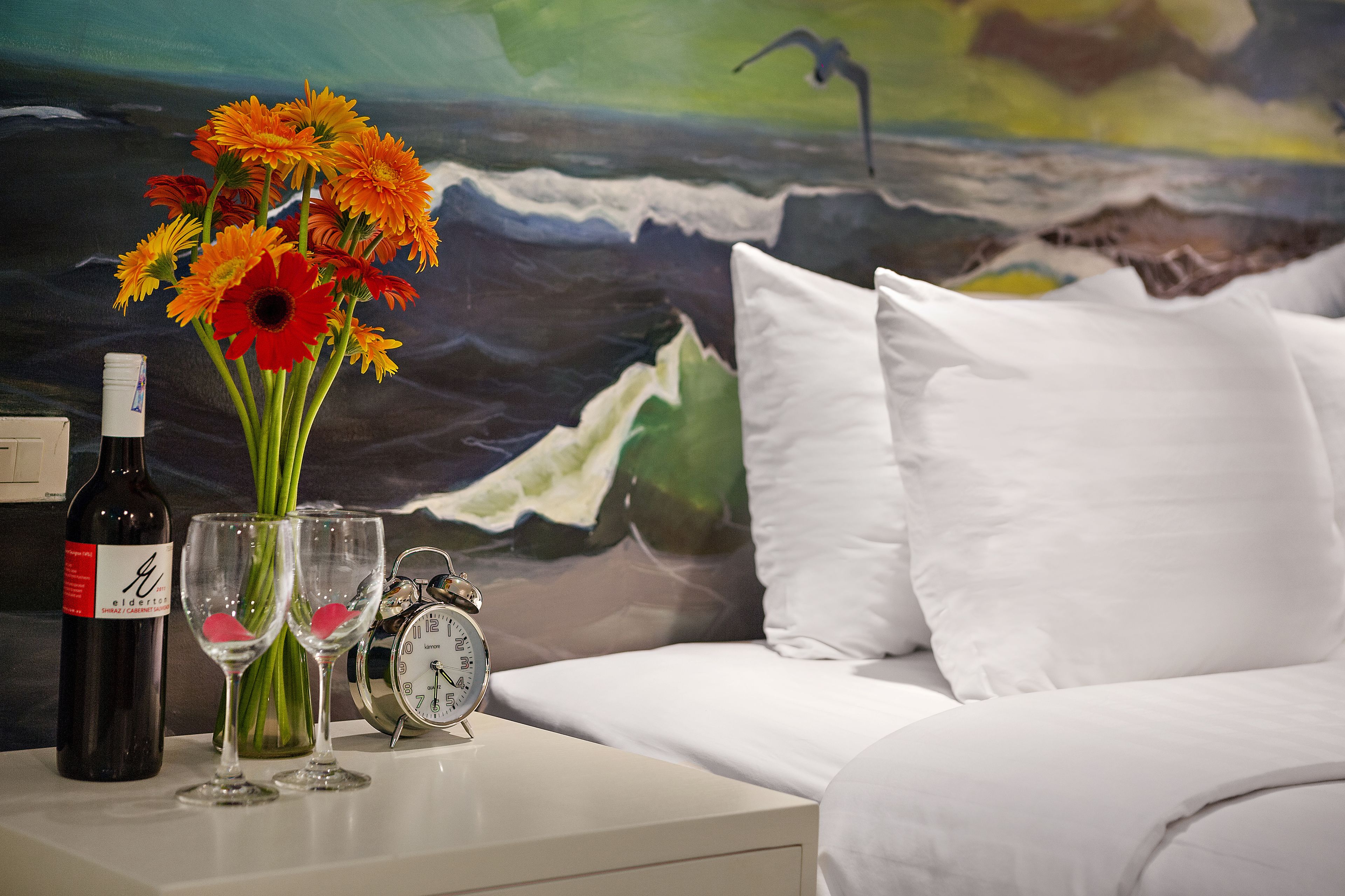 En seng med hvide puder, et hvidt natbord med en vinflaske, to vinglas og en vase med orange blomster.