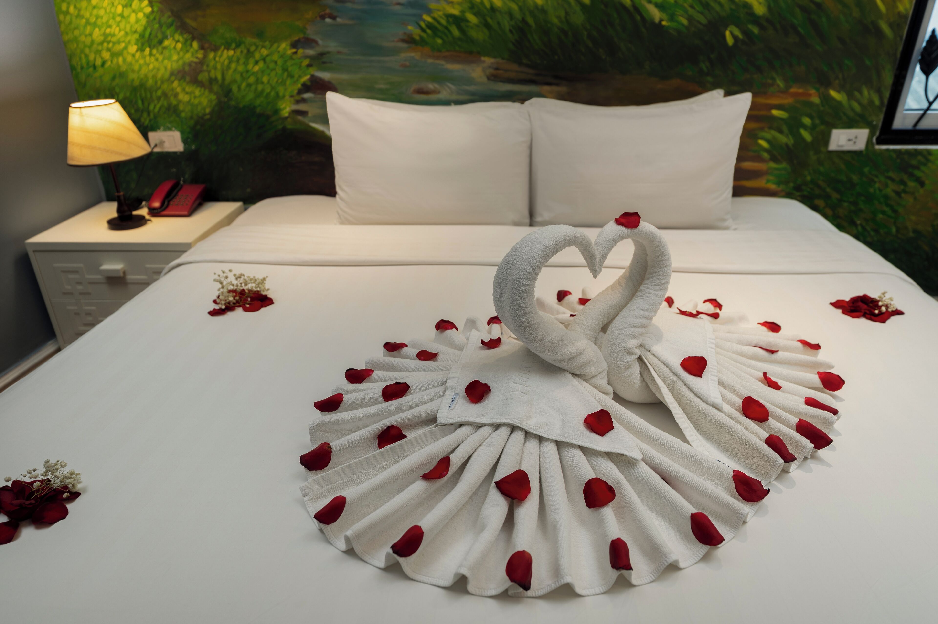 Una cama impecablemente hecha con un centro de toallas con forma de cisne, ropa de cama blanca y un arreglo de pétalos de rosa roja.