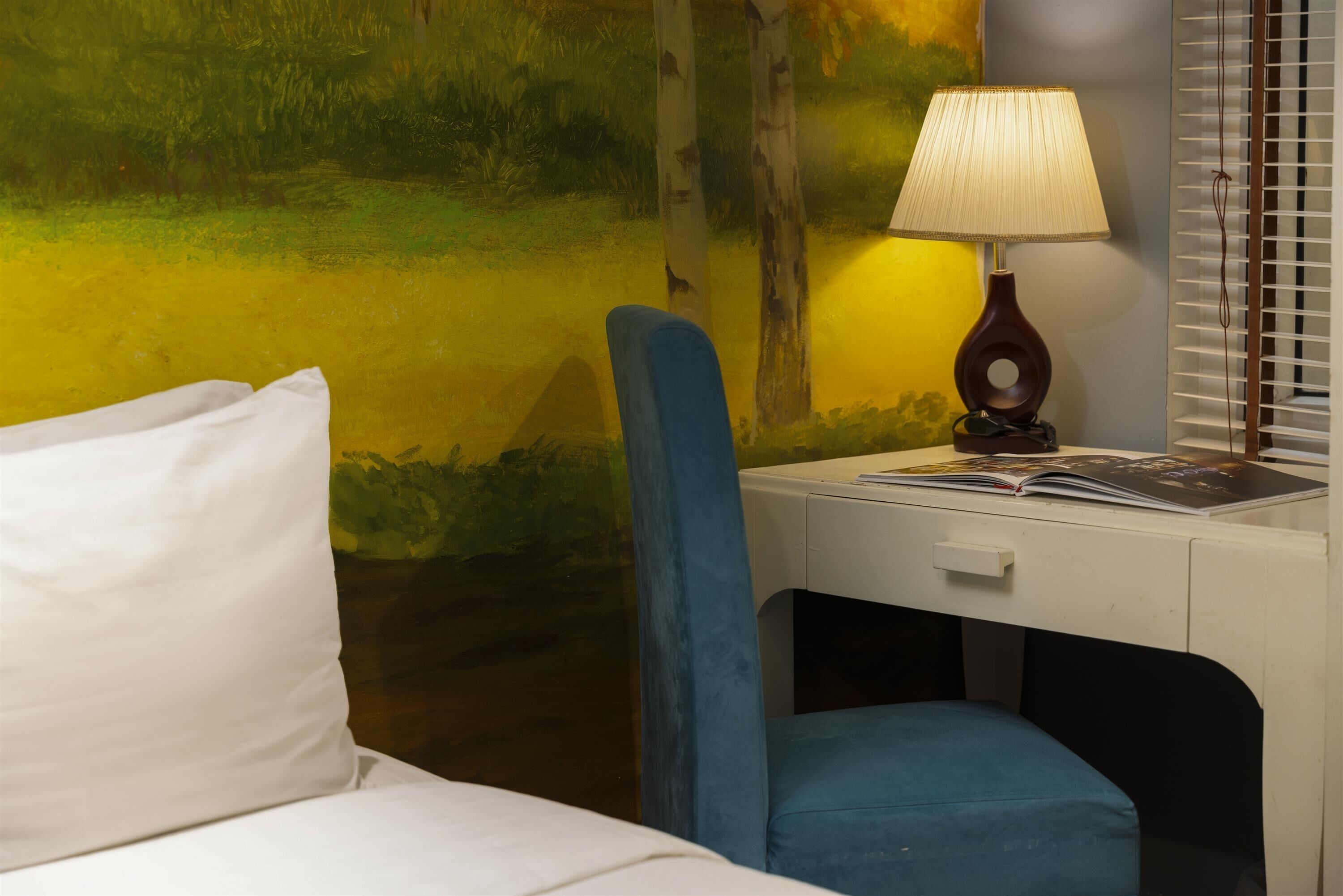 A hotel room with a bed, a desk with a lamp, and a wall with a painting.