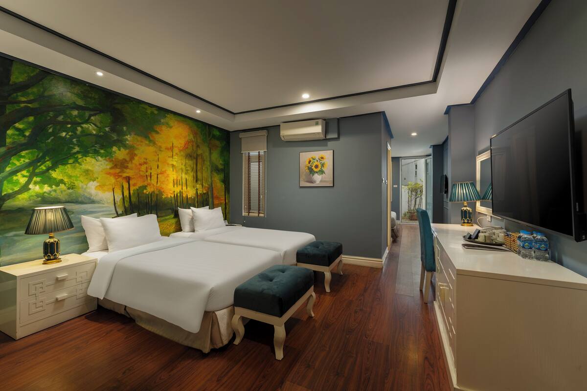 A modern bedroom with two beds, a large painting, a TV, and a small kitchenette.