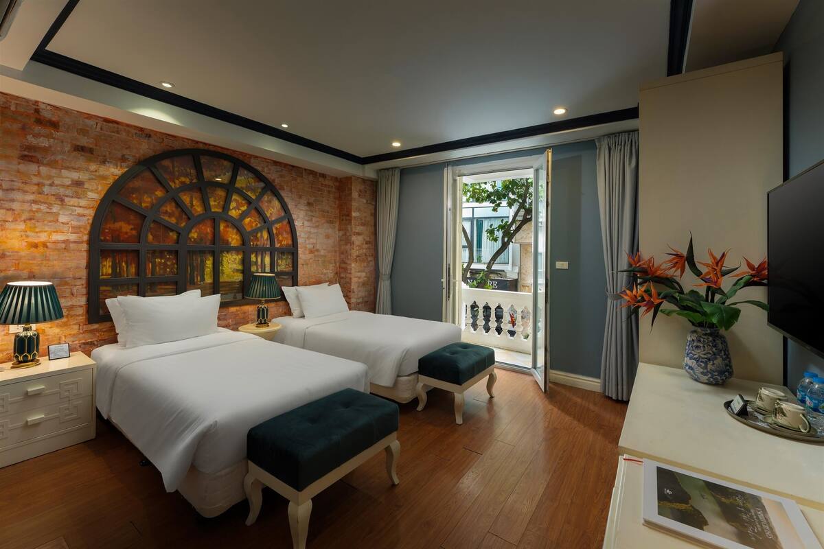 A hotel room with two beds, a brick wall, a large arched window, a balcony with a view, and a TV on a white table.