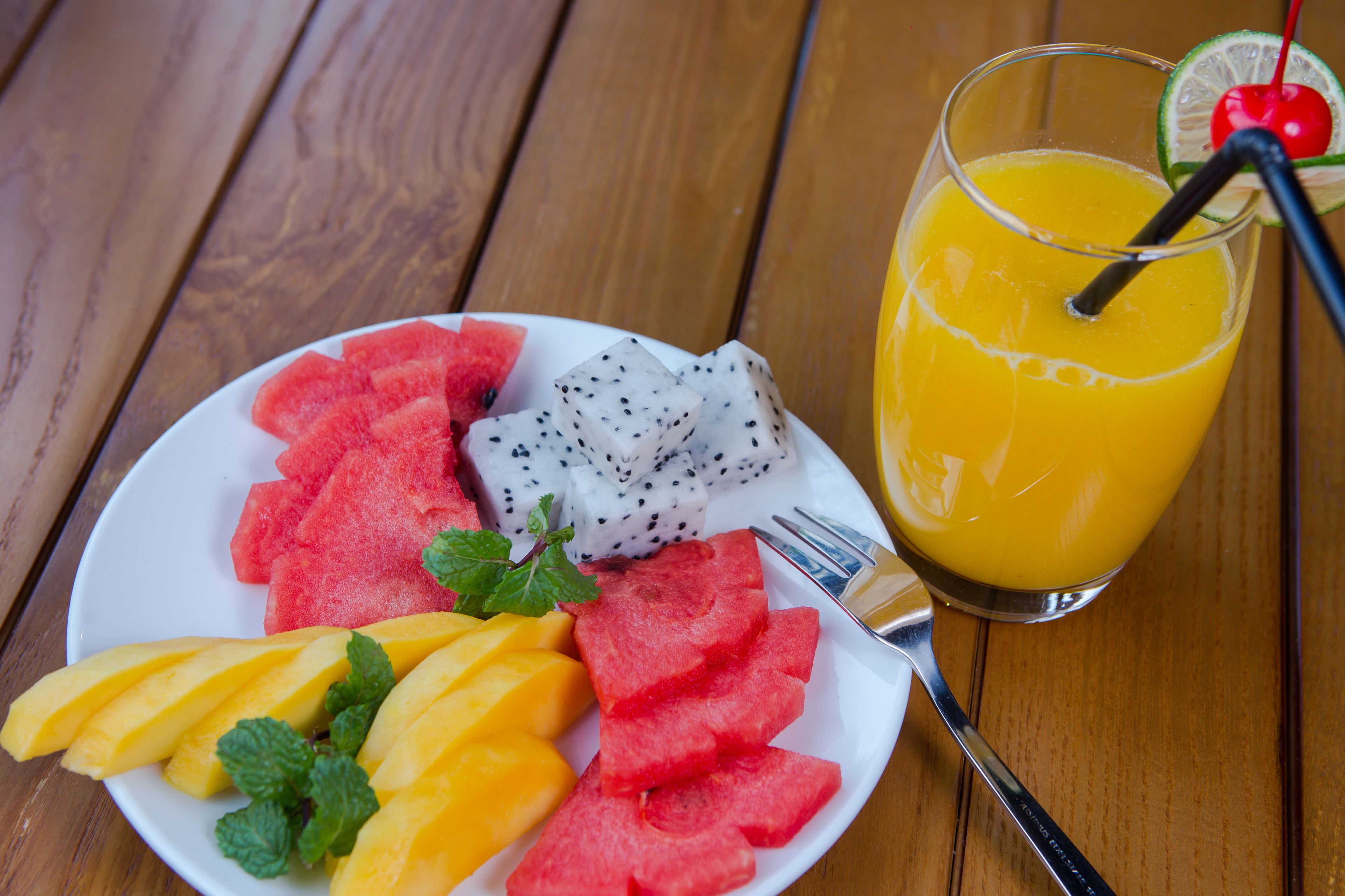 A plate of sliced watermelon, dragon fruit, and mango with a glass of orange juice and a cherry on the rim.