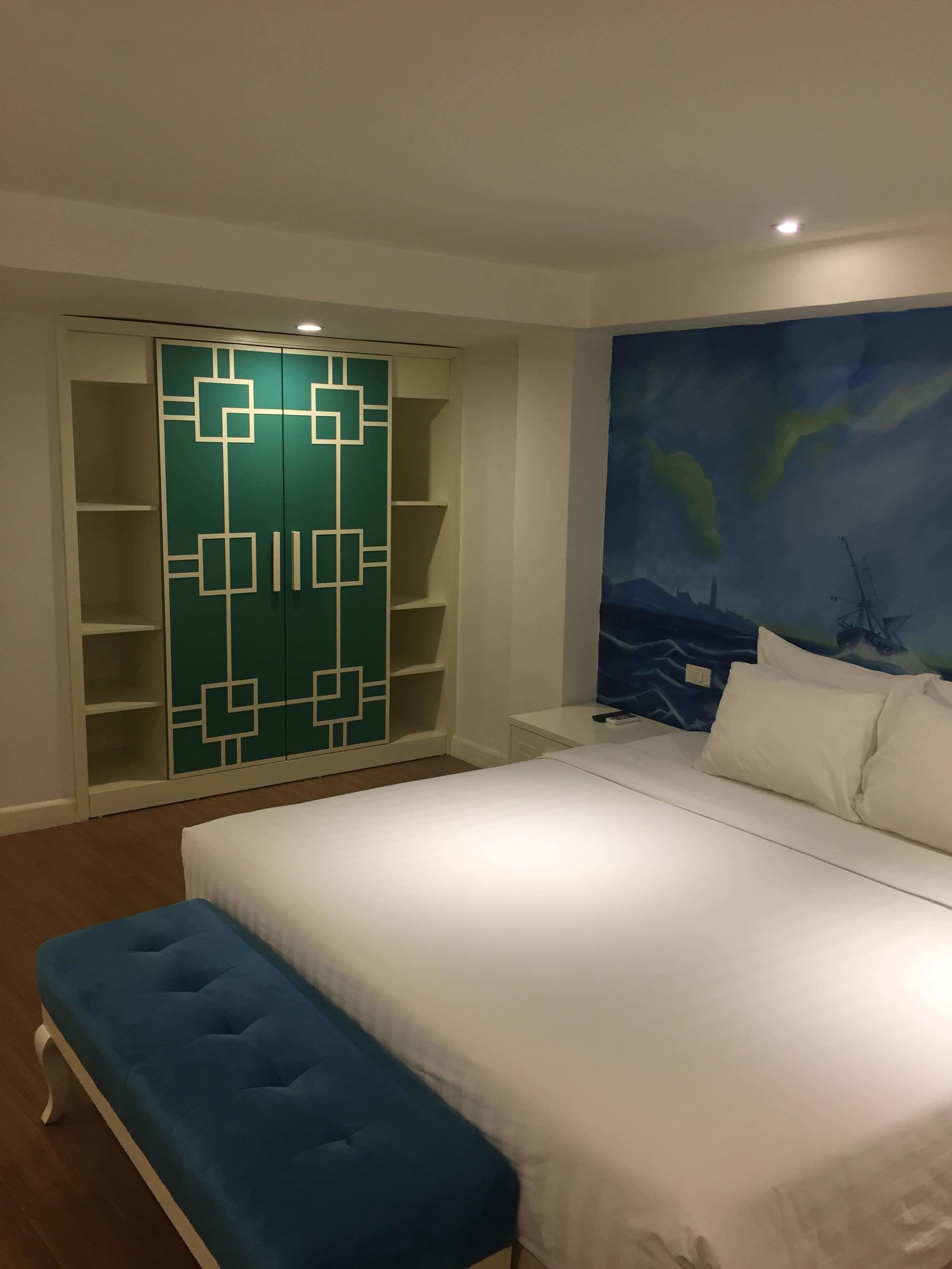 A bedroom with a bed, a blue bench, a wardrobe with geometric patterns, and a mural of a cityscape.