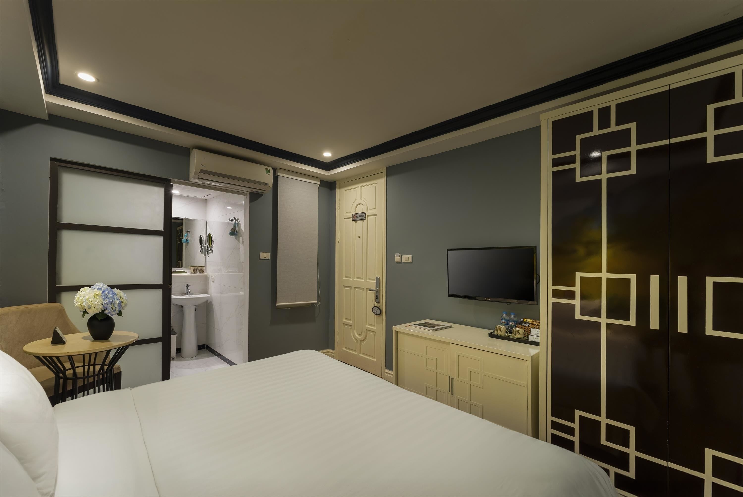 A hotel room with a bed, a desk with a chair, a TV, a wardrobe, and a bathroom visible through an open door.