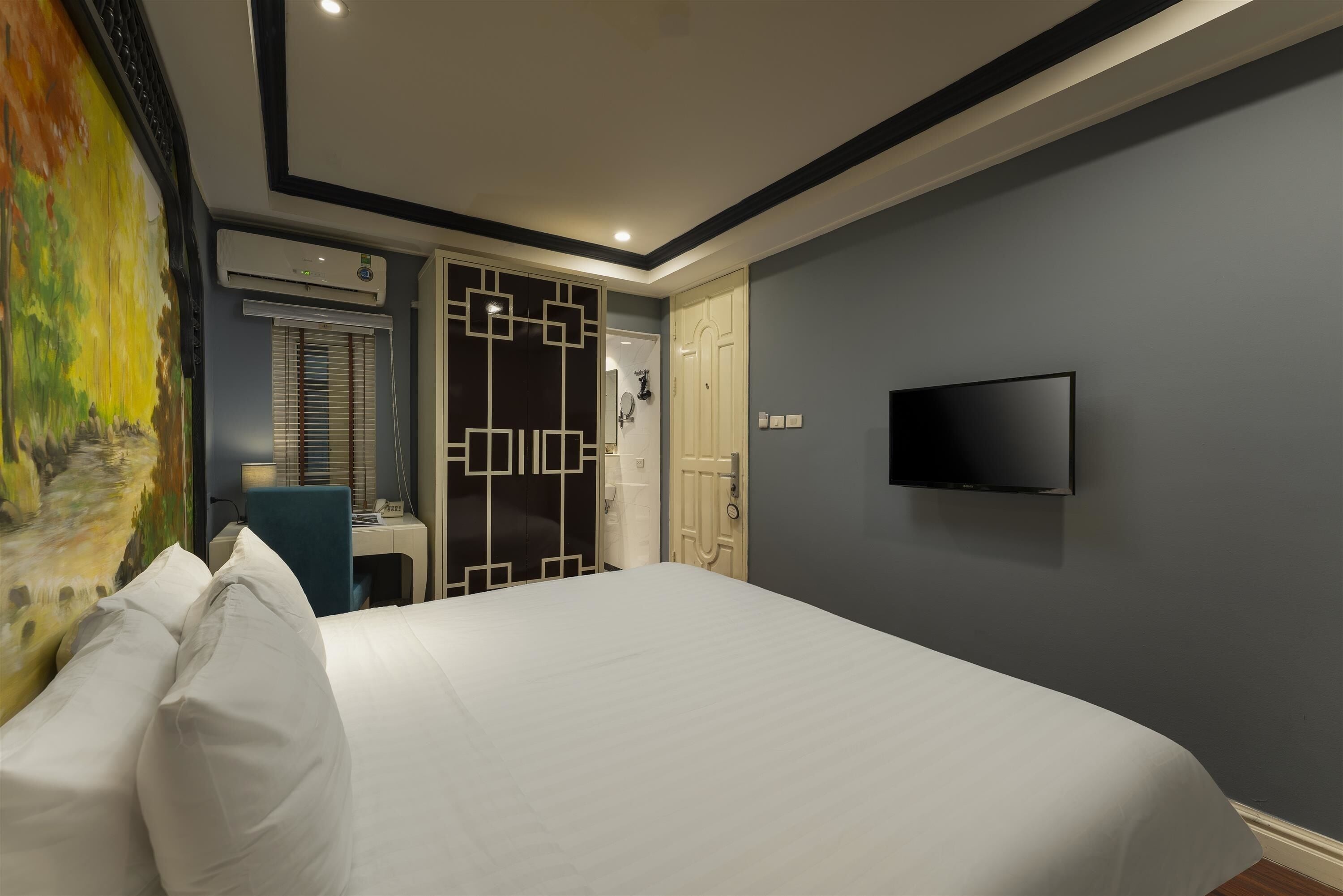 A hotel room with a bed, a TV mounted on the wall, an air conditioning unit, a door with a geometric design, and a painting on the wall.