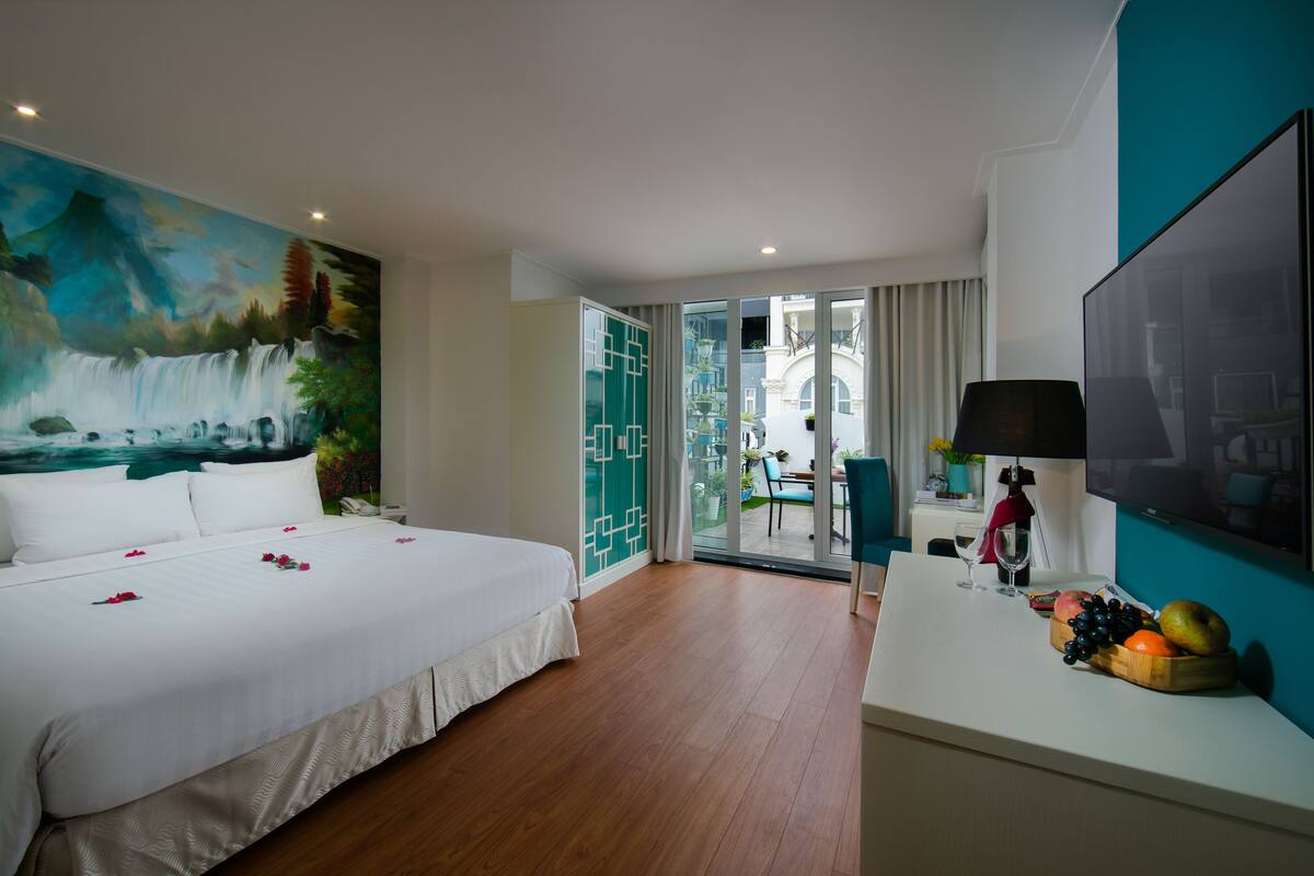 A hotel room with a large bed, a painting of a waterfall, a balcony with a view, and a fruit bowl on the nightstand.