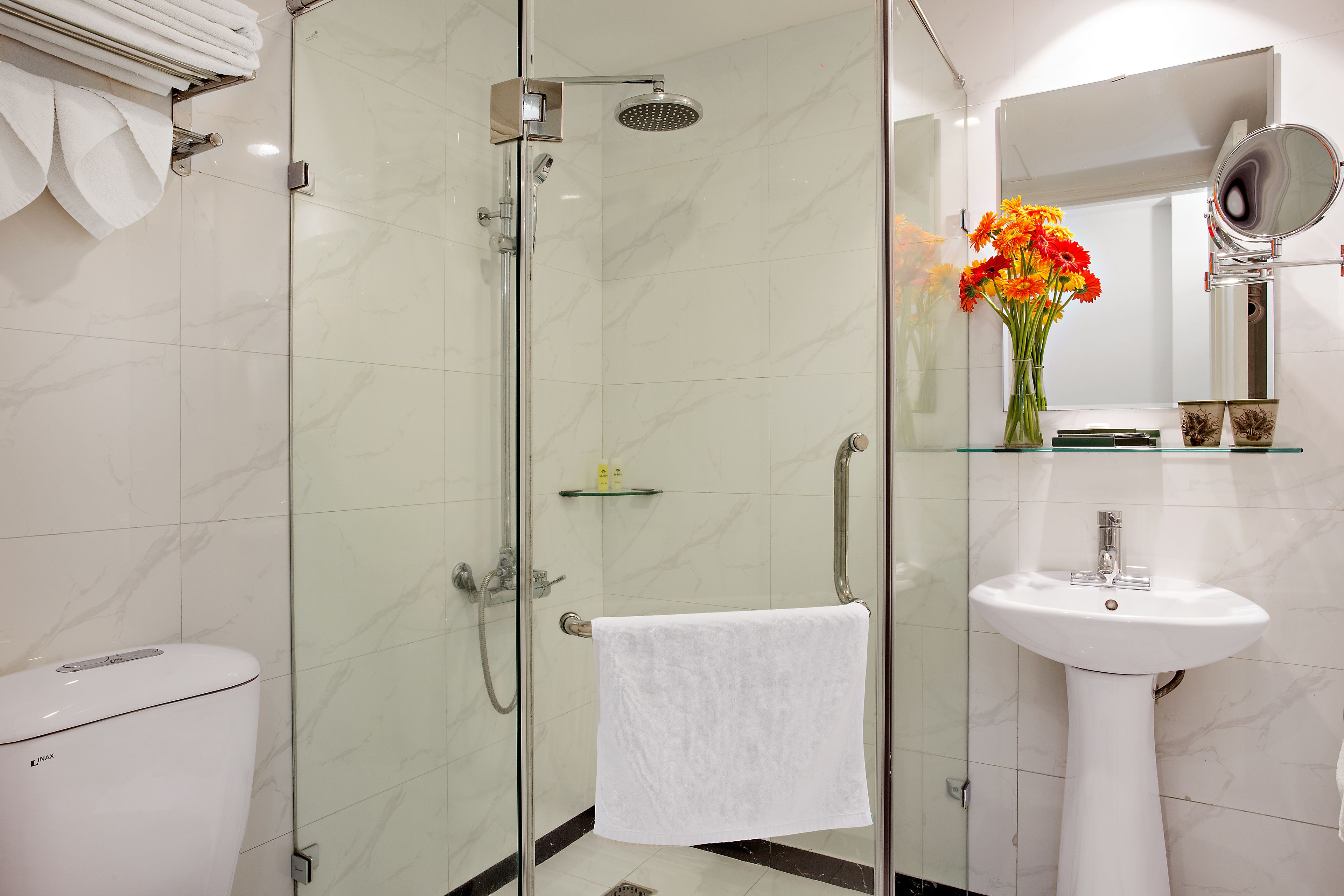 Un baño moderno con ducha de vidrio, inodoro blanco, lavabo con espejo y un jarrón con flores.