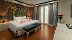A bedroom with a large bed, a bench, a nightstand, a lamp, a vase with flowers, and a view of the outdoors through a window.