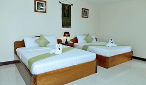 A hotel room with two single beds, each with a wooden headboard, white bedding, and green throw pillows. There is a nightstand with a lamp and a small table between the beds.