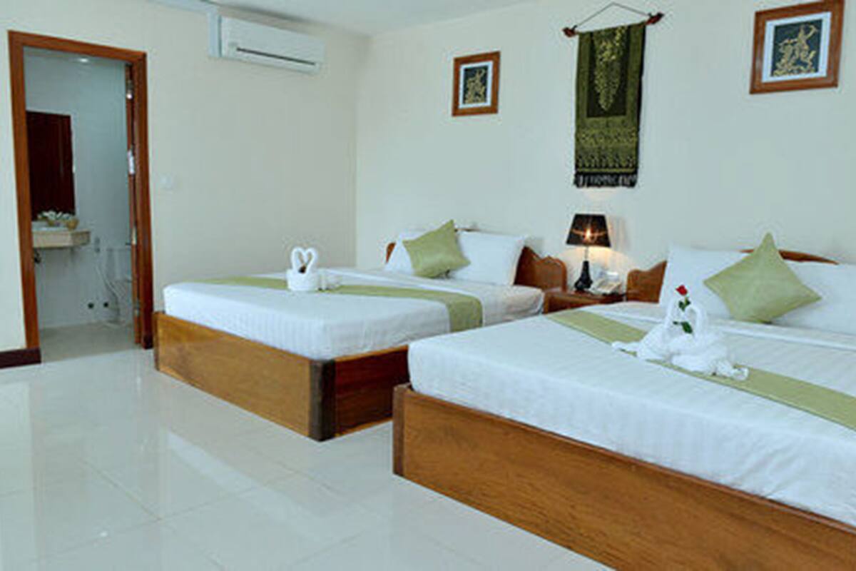 A hotel room with two beds, each with a wooden frame, white bedding, and green pillows. There is a small table with a lamp, a picture on the wall, and an air conditioning unit on the wall.