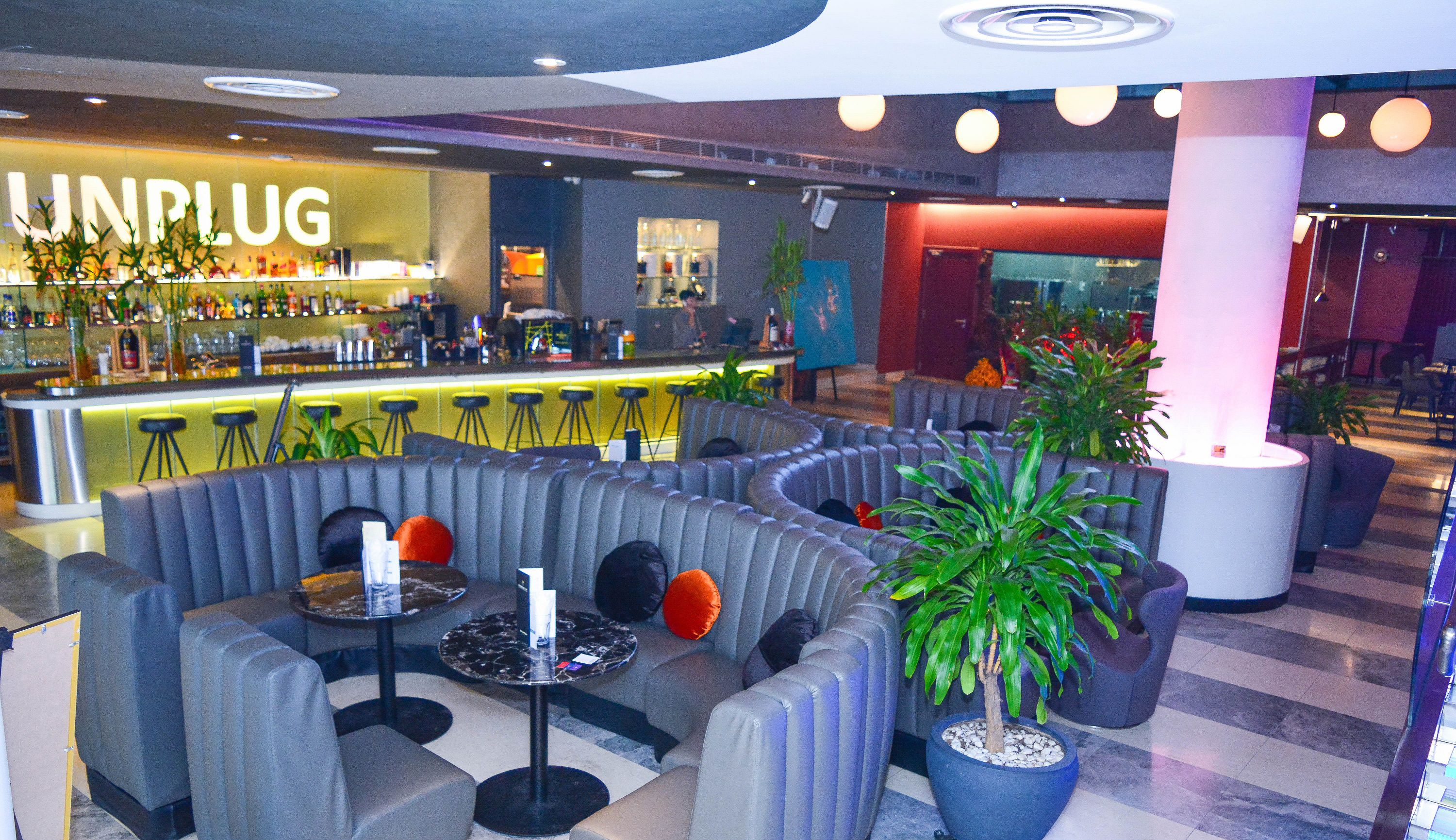 A modern lounge area with a bar named "UNPLUG" in the background, featuring comfortable seating and a variety of plants.