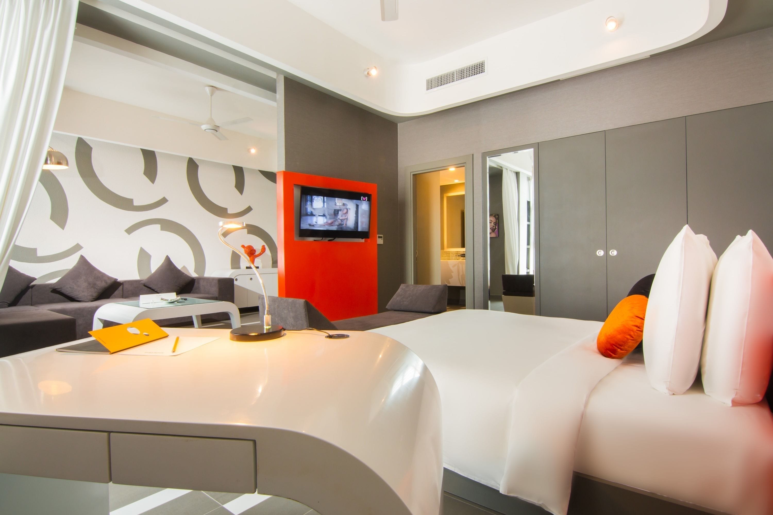 A modern hotel room with a bed, a sofa, a TV, and a desk with a lamp.