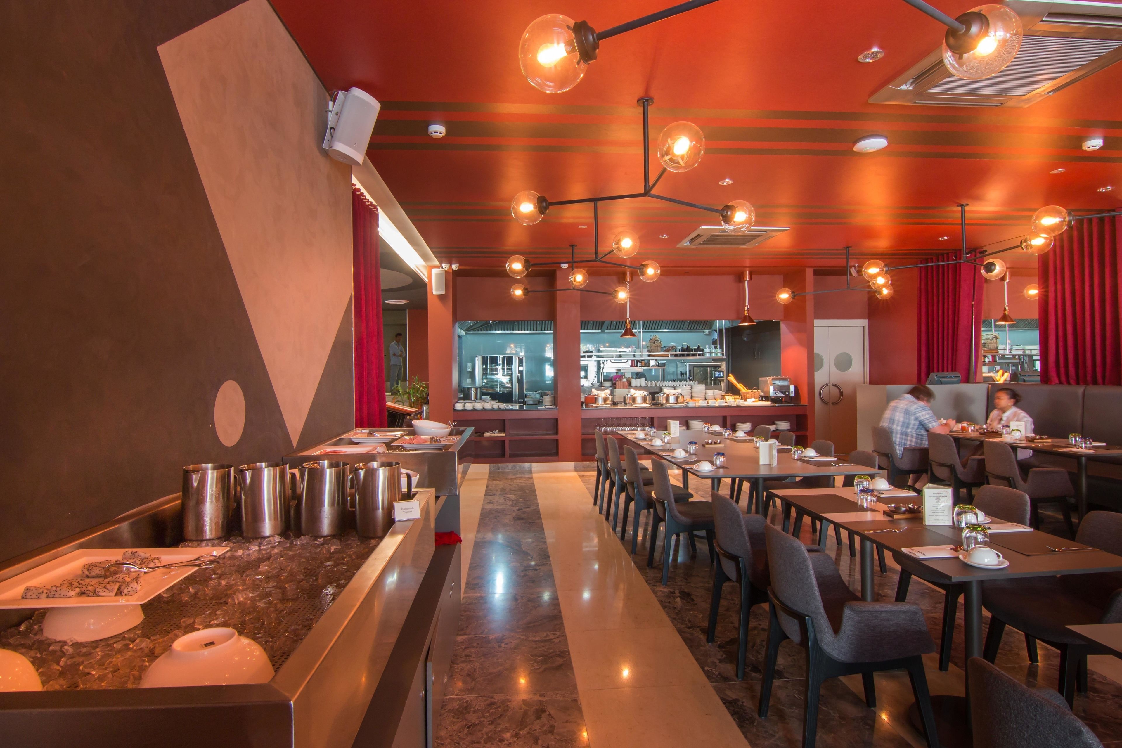 A restaurant with a modern interior, featuring a long counter with a variety of dishes, multiple dining tables, and a bar area in the background.
