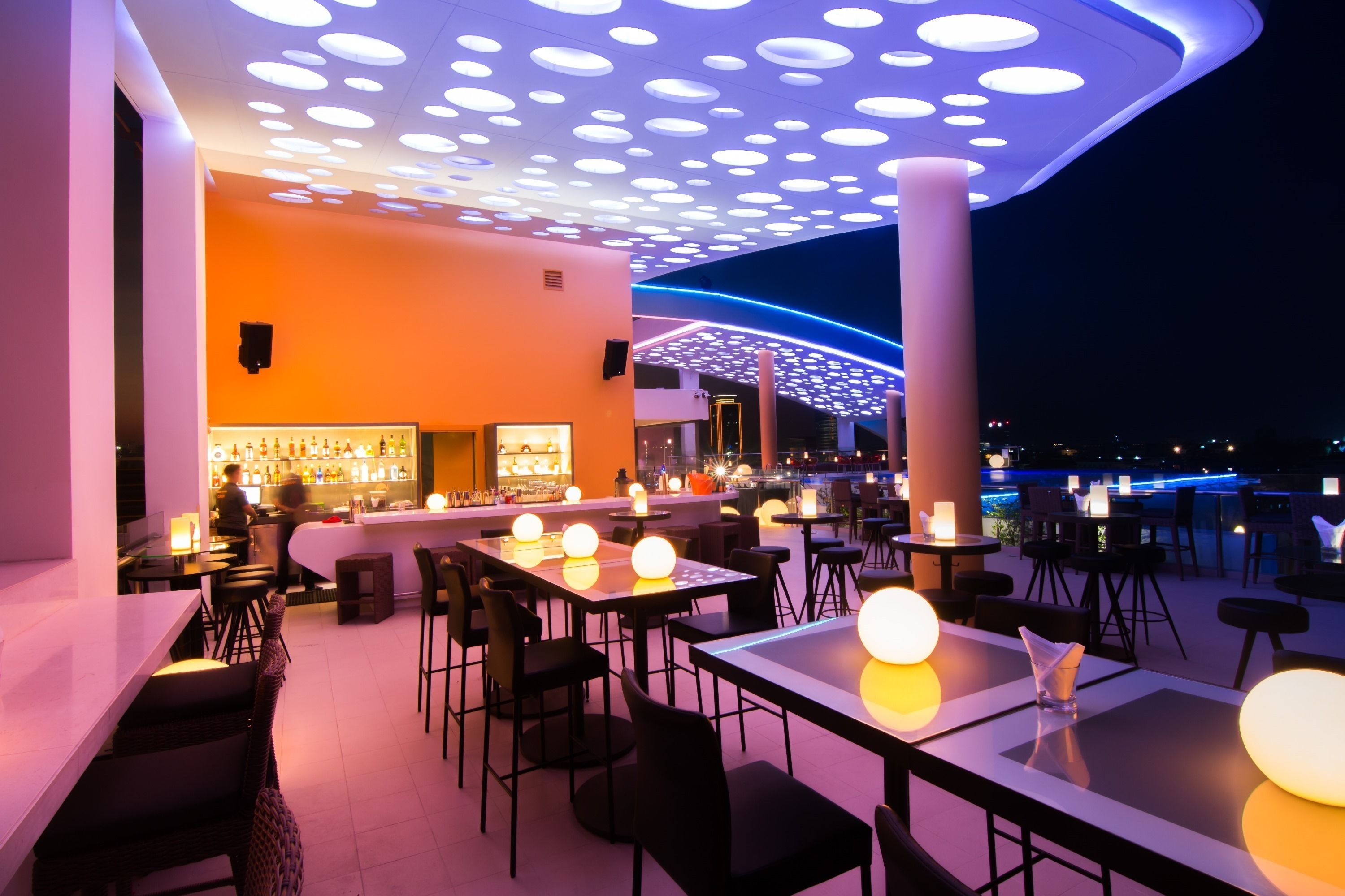 A modern restaurant with a bar area, round tables, and a ceiling with circular light fixtures.