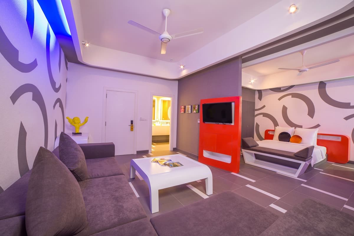 A modern hotel room with a sofa, a white coffee table, a red bed, and a TV.