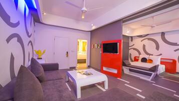 Una habitación de hotel moderna con sofá, una mesita de café blanca, una cama roja y un televisor.