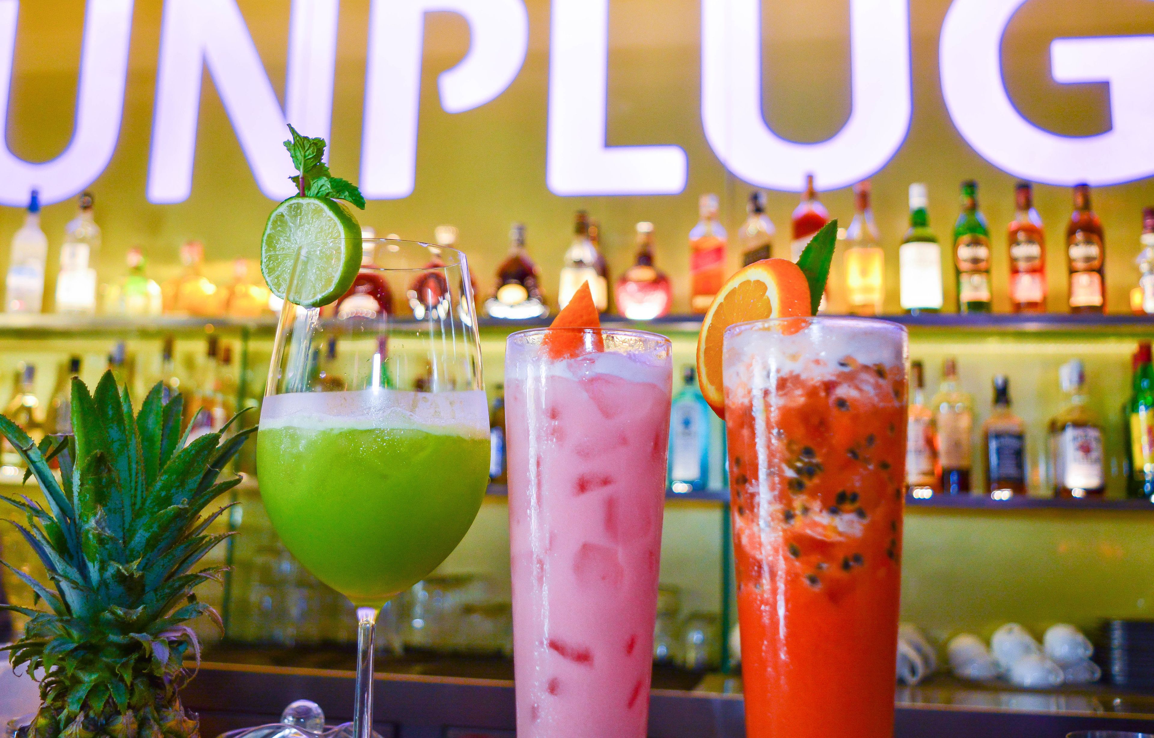 Trois cocktails décorés, un ananas et un bar avec différentes bouteilles en arrière-plan.