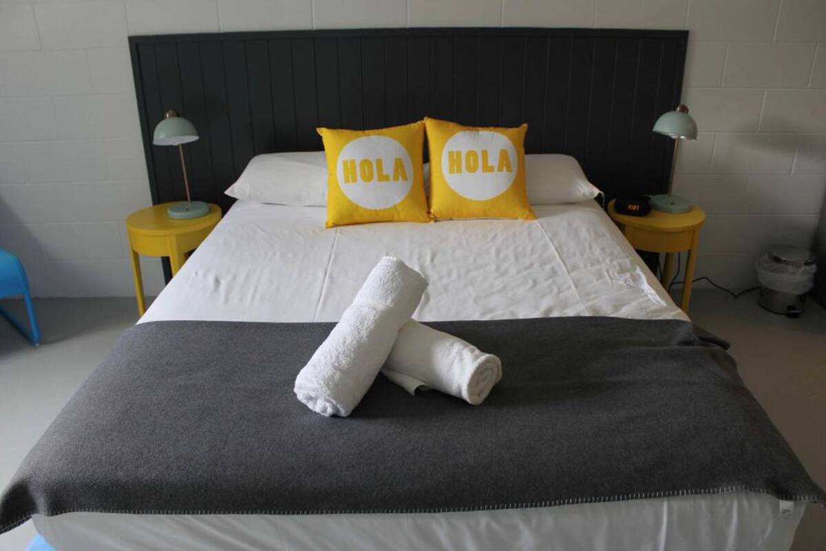 Una cama bien hecha con sábanas blancas, dos almohadas con el texto "HOLA", una toalla blanca enrollada y dos lámparas de noche sobre mesitas amarillas.