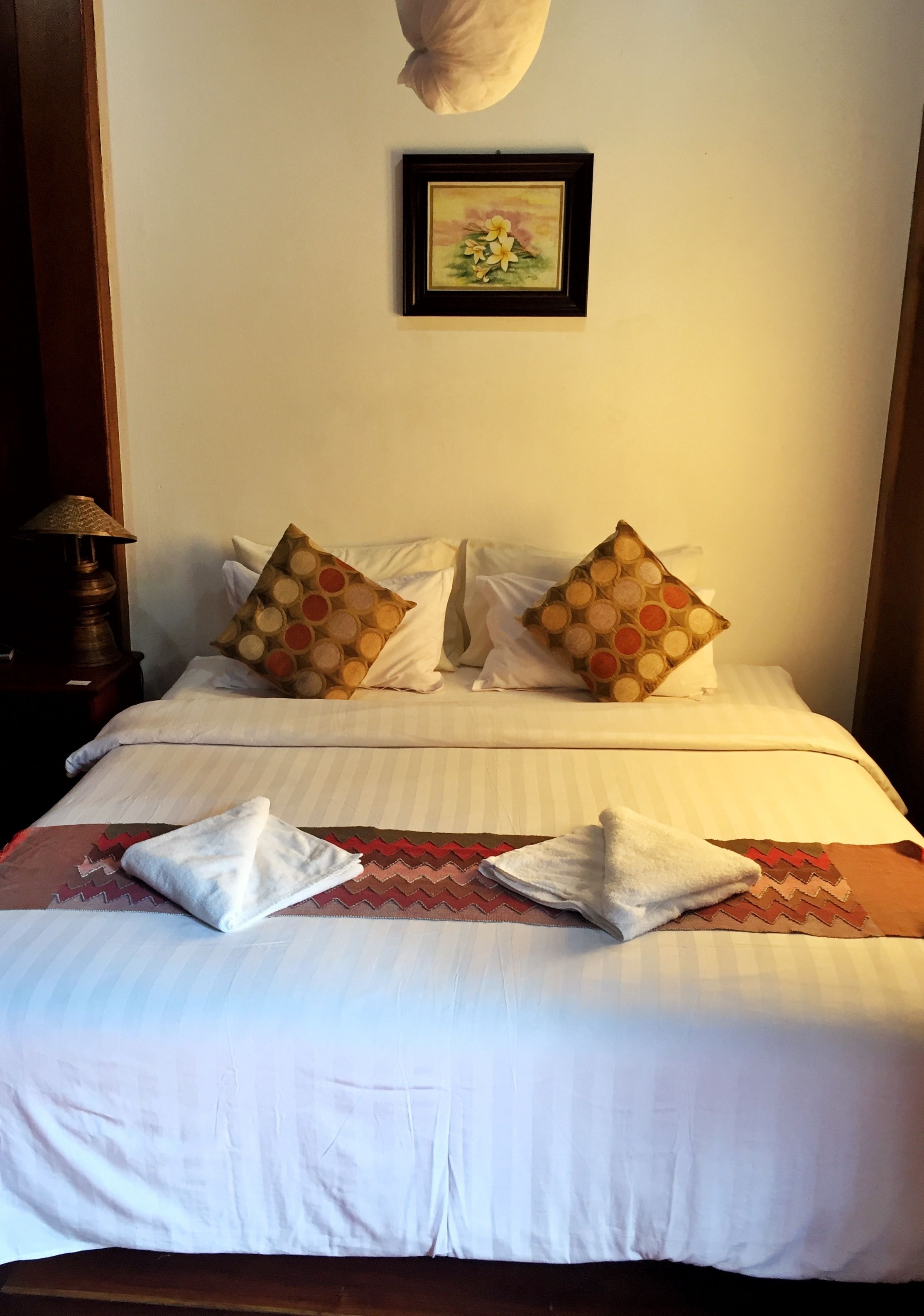Una cama bien hecha con almohadones estampados, un cuadro en la pared y una lámpara en la mesita de noche.