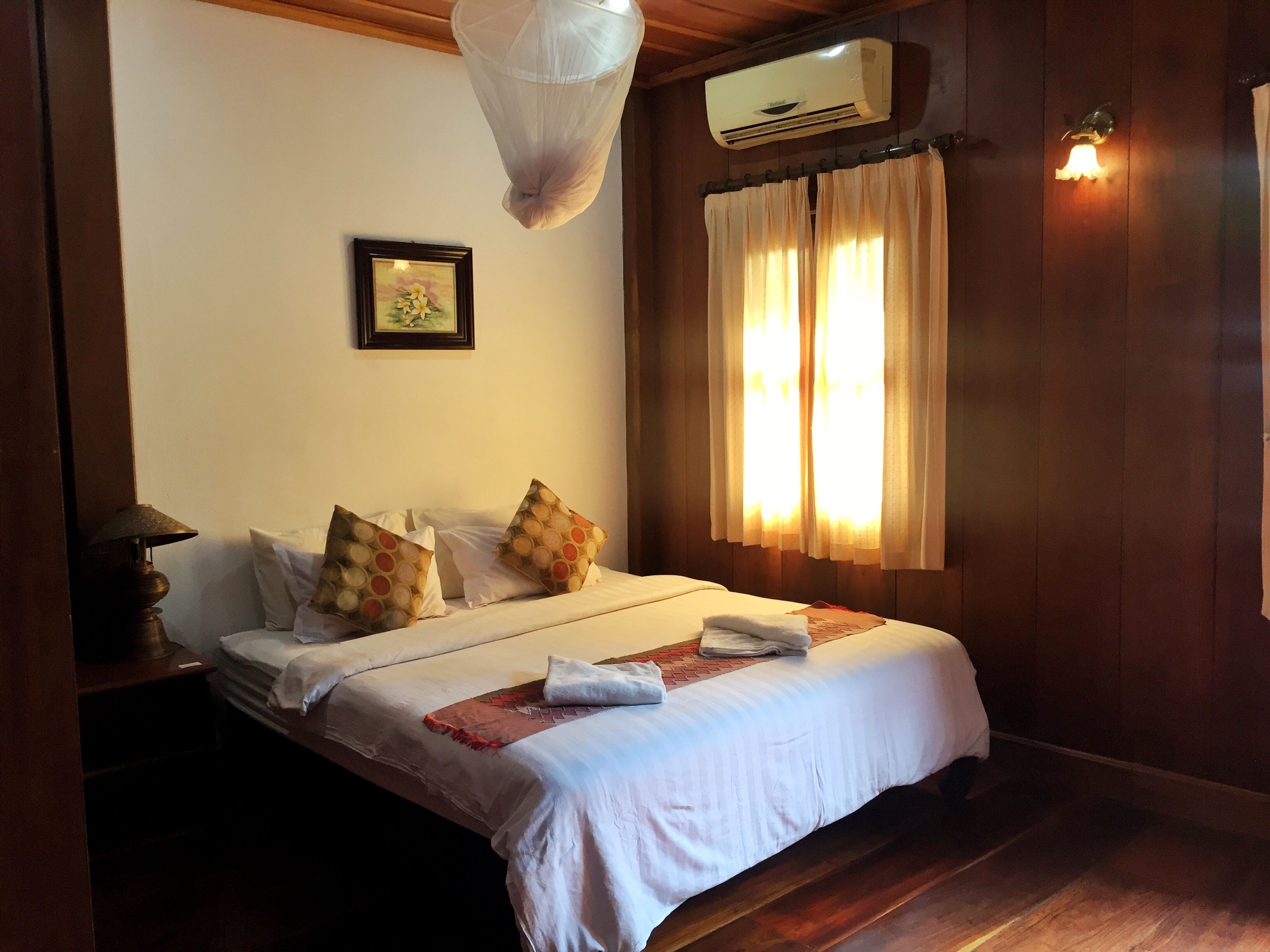 A neatly made bed with white linens and decorative pillows, a bedside lamp, and a wall-mounted air conditioner.